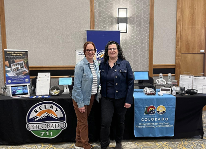Two women standing in front of an exhibition table with Relay Colorado 711 banner and Colorado Commission for the Deaf, Hard of Hearing, and DeafBlind materials at conference or expo